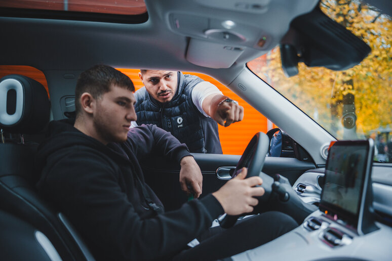 Ein junger Mann sitzt mit den Händen am Lenkrad auf dem Fahrersitz eines Autos, während sich ein anderer Mann außerhalb des Autos durch das Fenster beugt, zeigt und Anweisungen gibt. Im Hintergrund sind eine orangefarbene Wand und Herbstbäume zu sehen.