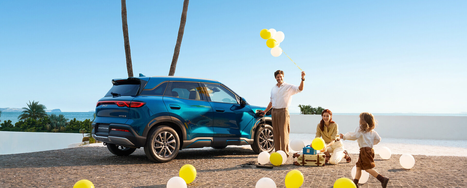 Familie mit zwei Erwachsenen und einem Kind spielt mit gelben und weißen Luftballons neben einem blauen SUV auf einem sandigen Platz mit Palmen im Hintergrund