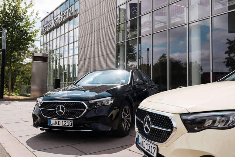 Zwei Mercedes-Benz Fahrzeuge, ein schwarzer und ein beiger, stehen vor einem modernen Gebäude mit Glasfassade. Die Fahrzeuge zeigen das Markenlogo auf der Kühlerhaube.