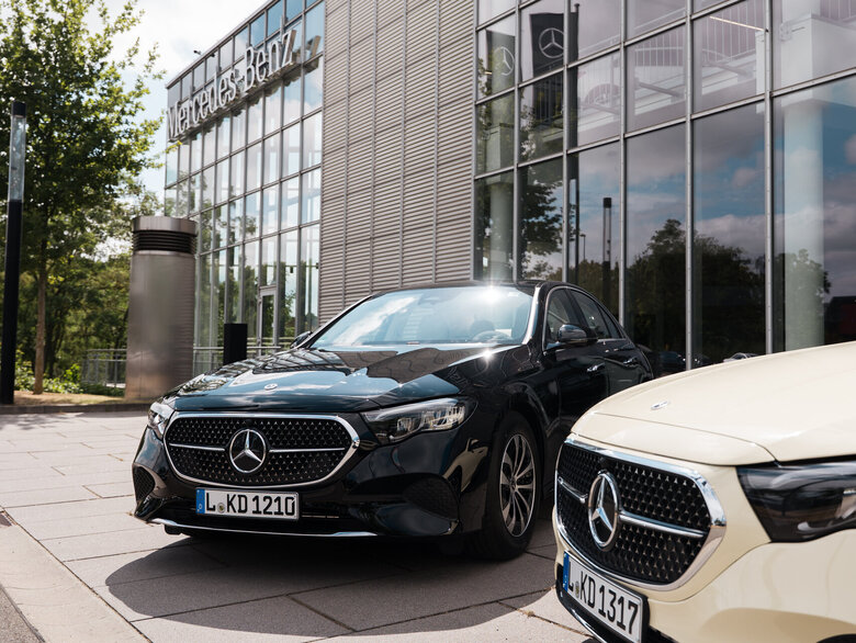 Zwei Mercedes-Benz Fahrzeuge, ein schwarzer und ein beiger, stehen vor einem modernen Gebäude mit Glasfassade. Die Fahrzeuge zeigen das Markenlogo auf der Kühlerhaube.