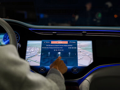 Die Hand einer Person interagiert mit einem Touchscreen auf dem Armaturenbrett eines Autos, auf dem Navigations- und Ladestationsoptionen angezeigt werden, mit beleuchteten digitalen Bedienelementen und einer Karte im Hintergrund.