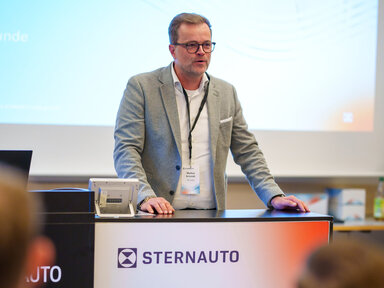 Markus Schmidt im grauen Anzug steht an einem Podium mit dem STERNAUTO-Logo und spricht zu einem Publikum in einem Konferenzraum. Hinter ihm ist ein Präsentationsbildschirm zu sehen.