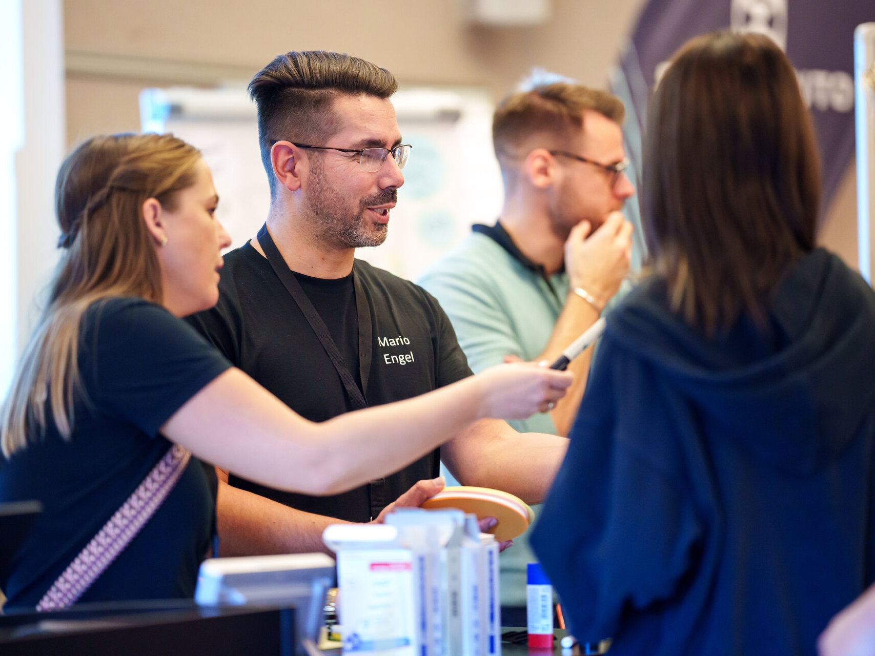 Ein Mann und zwei Frauen unterhalten sich an einem Stand. Auf der Theke liegen verschiedene kleine Produkte. Ein weiterer Mann steht im Hintergrund, teilweise unscharf.