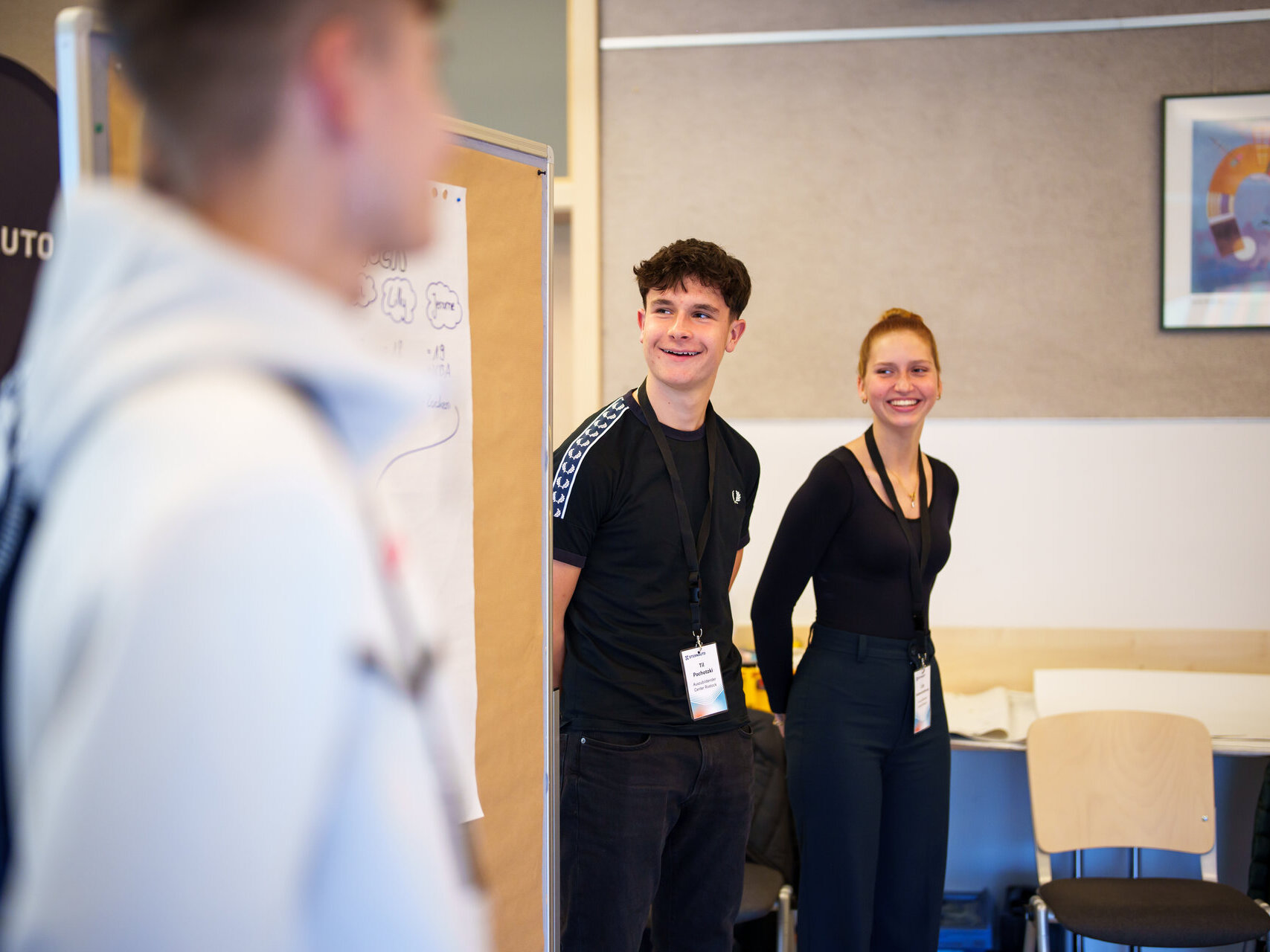 Zwei junge Erwachsene stehen lächelnd neben einer Tafel mit Zeichnungen, während eine weitere Person im Vordergrund steht. Sie befinden sich in einem Klassenzimmer oder einer Werkstatt und tragen Namensschilder.