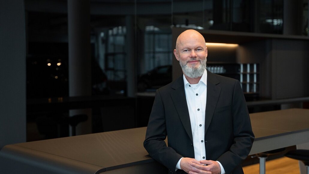 Ein kahlköpfiger Mann (Peter Heinrich) mit grauem Bart, schwarzem Anzug und weißem Hemd, steht in einem modernen Büro mit dunklen Wänden und Glasfenstern und lächelt.