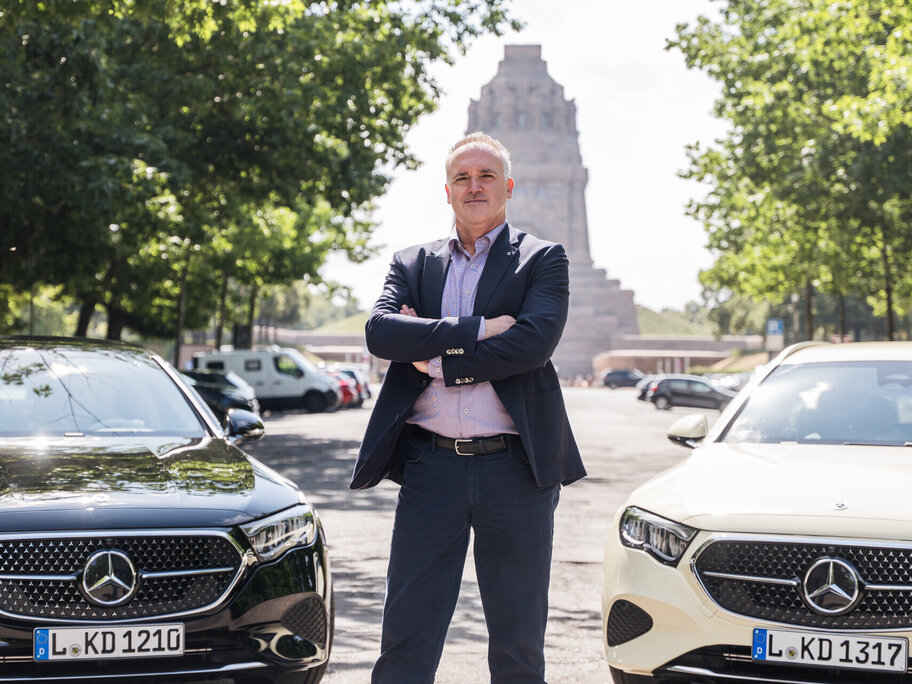 Ein Mann steht mit verschränkten Armen zwischen zwei Mercedes-Benz Autos, einem schwarzen und einem weißen, vor einem monumentalen Gebäude.