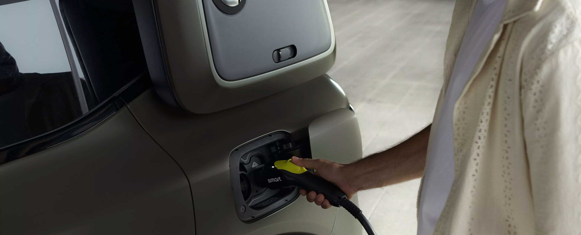 Ein Mann schließt ein Ladekabel an ein kleines Elektroauto an. Der Fokus liegt auf dem Ladeanschluss des Autos und der Hand des Mannes, die das Kabel hält. Die Szene ist gut beleuchtet und der Hintergrund ist unscharf.