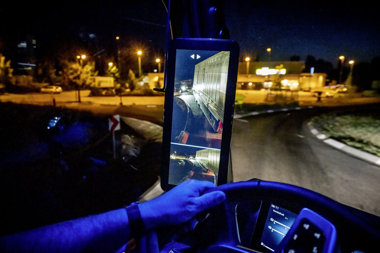 19C0843_19 Eine Person fährt einen Lkw bei Nacht und hält das Lenkrad mit einer Hand. Ein digitales Rückfahrdisplay zeigt Seitenansichten des Lastwagens und der Straße, während draußen Straßenlaternen und Gebäude zu sehen sind.