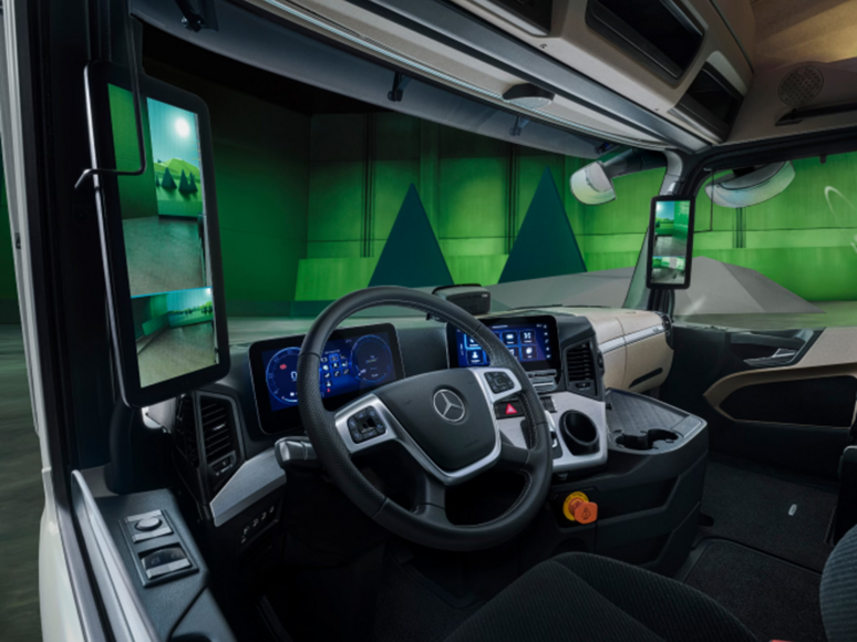 Innenansicht einer modernen Mercedes-Benz Lkw-Kabine mit digitalem Armaturenbrett, Lenkrad und zwei großen digitalen Seitenspiegeln, wobei außerhalb der Fenster eine grün beleuchtete Industrieumgebung zu sehen ist.