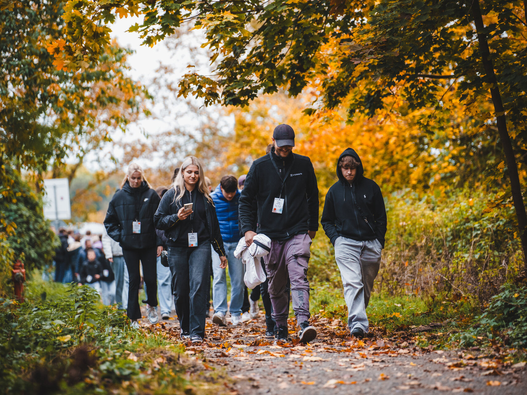Eine Gruppe von Menschen geht in Jacken und mit Schlüsselbändern bekleidet einen blätterbedeckten Weg in einem von Herbstbäumen umgebenen Park entlang. Im Hintergrund sind weitere Teilnehmer zu sehen.
