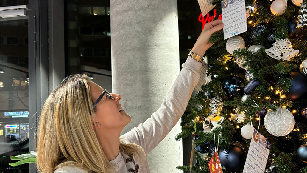 Eine Frau mit Brille und Rentierpulli lächelt, als sie eine Karte an einen geschmückten Weihnachtsbaum mit Lichtern, Ornamenten und Wunschkarten hängt.