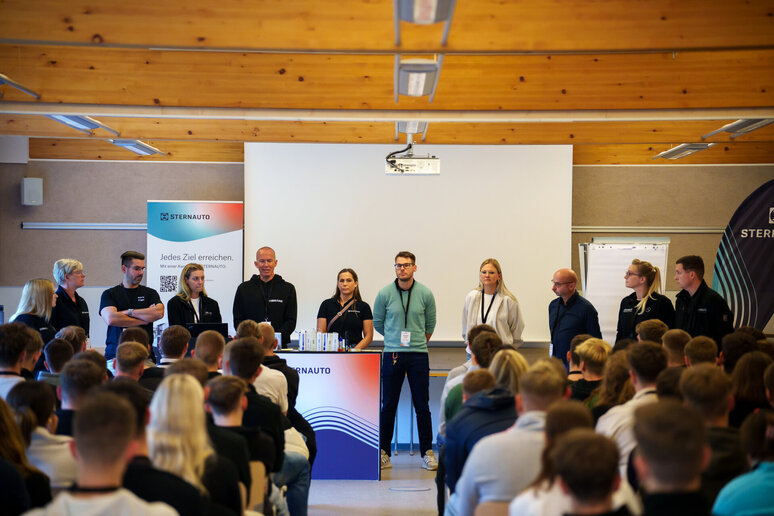 Eine Gruppe von Rednern steht in einem Klassenzimmer oder Seminarraum vor einem sitzenden Publikum und hält eine Rede. Hinter ihnen sind ein Präsentationsbildschirm und Banner zu sehen.