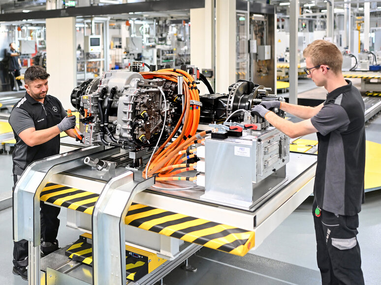 Zwei Arbeiter in einer Fabrik montieren einen Elektromotor mit sichtbaren orangefarbenen Kabeln an einem Arbeitsplatz, umgeben von Industrieanlagen und Maschinen in einer hellen, modernen Anlage.