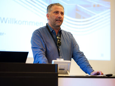 Ein Mann steht und spricht an einem Podium mit der Aufschrift STERNAUTO in einem modernen Konferenzraum. Im Hintergrund ist eine Präsentationsfolie mit dem Firmenlogo und dem Wort „Willkommen“ zu sehen.