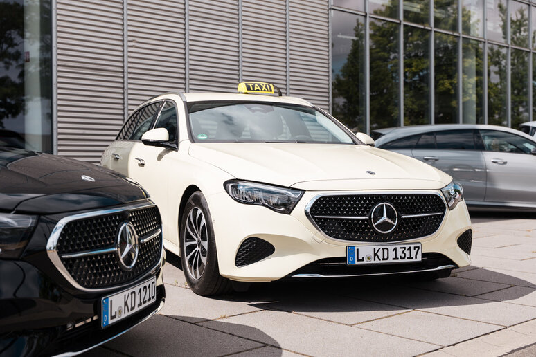 Ein weißer Mercedes-Benz mit Taxischild auf dem Dach steht neben einem schwarzen Mercedes-Benz. Beide Fahrzeuge sind auf einem Parkplatz.