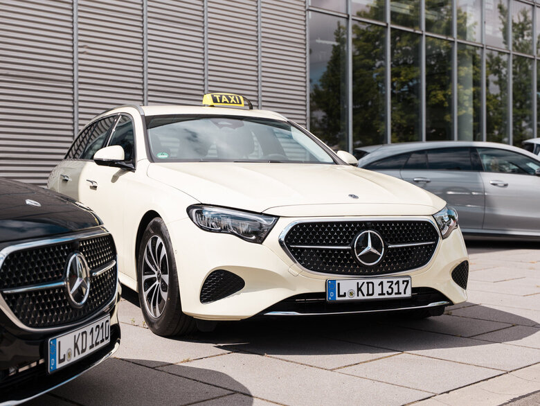 Ein weißer Mercedes-Benz mit Taxischild auf dem Dach steht neben einem schwarzen Mercedes-Benz. Beide Fahrzeuge sind auf einem Parkplatz.