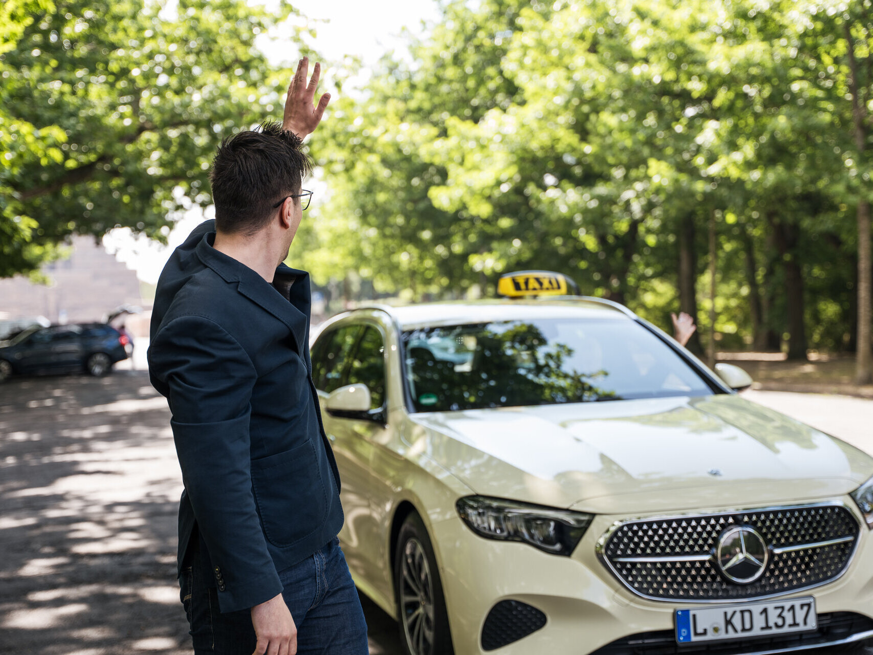 Ein Mann in einem dunkelblauen Anzug winkt einem weißen Taxi mit einem gelben Dachzeichen zu. Bäume sind im Hintergrund sichtbar.