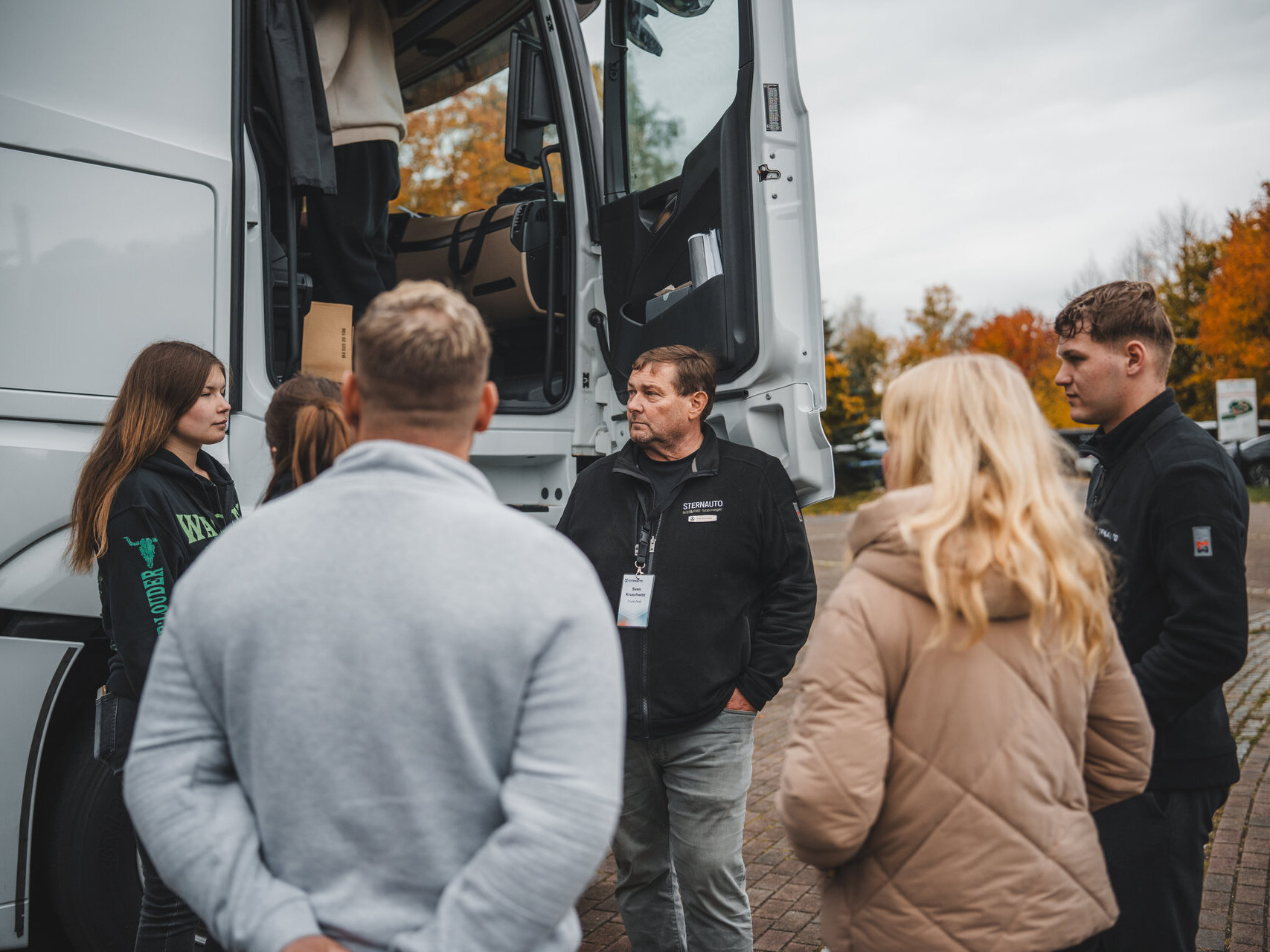 Eine Gruppe von Personen steht um einen Lastwagen und hört einem Mann zu, der Informationen gibt. Die Personen sind in lässiger Kleidung gekleidet und zeigen Interesse. Bunte Herbstbäume sind im Hintergrund sichtbar.