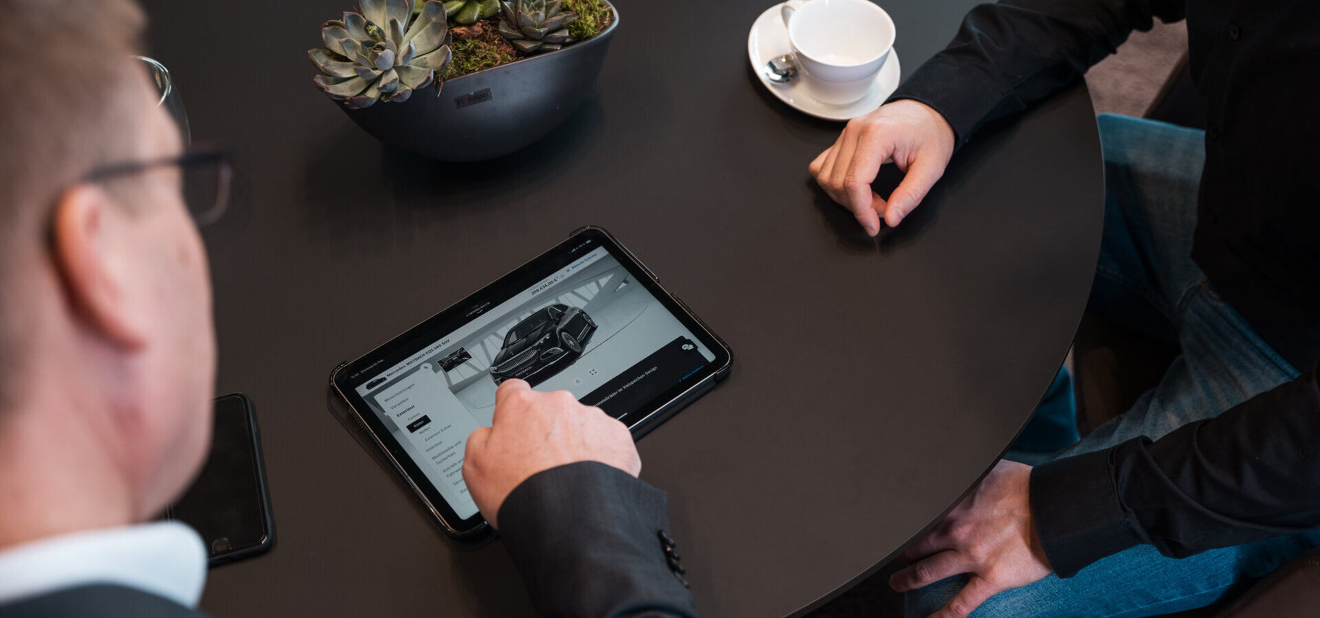 Drei Menschen bei einer Fahrzeugberatung - Ein Mann zeigt auf ein Fahrzeugangebot auf einem Tablet