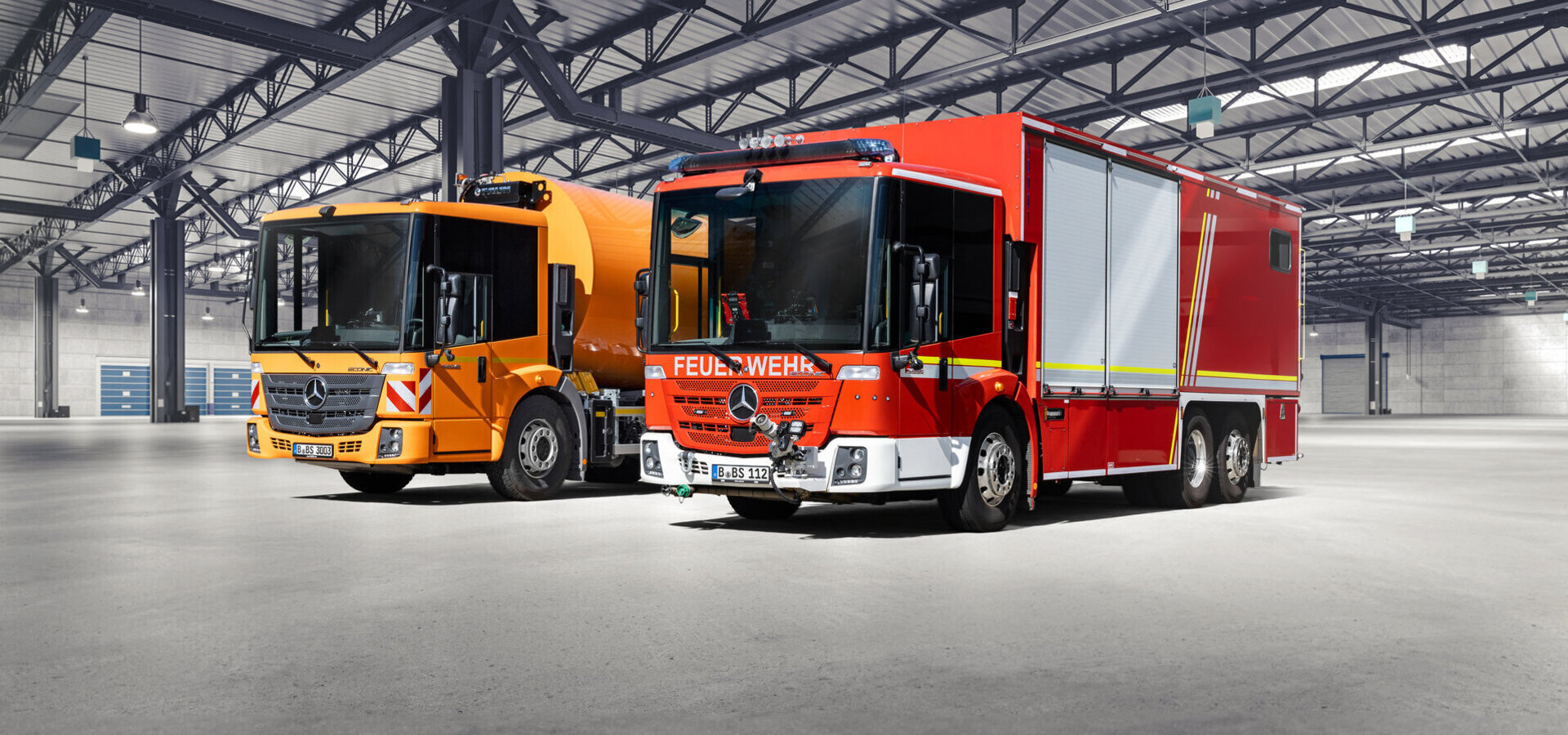 Zwei Einsatzfahrzeuge, ein oranger Lkw und ein rotes Feuerwehrfahrzeug, stehen in einer großen Halle mit grauem Boden und weißer Decke