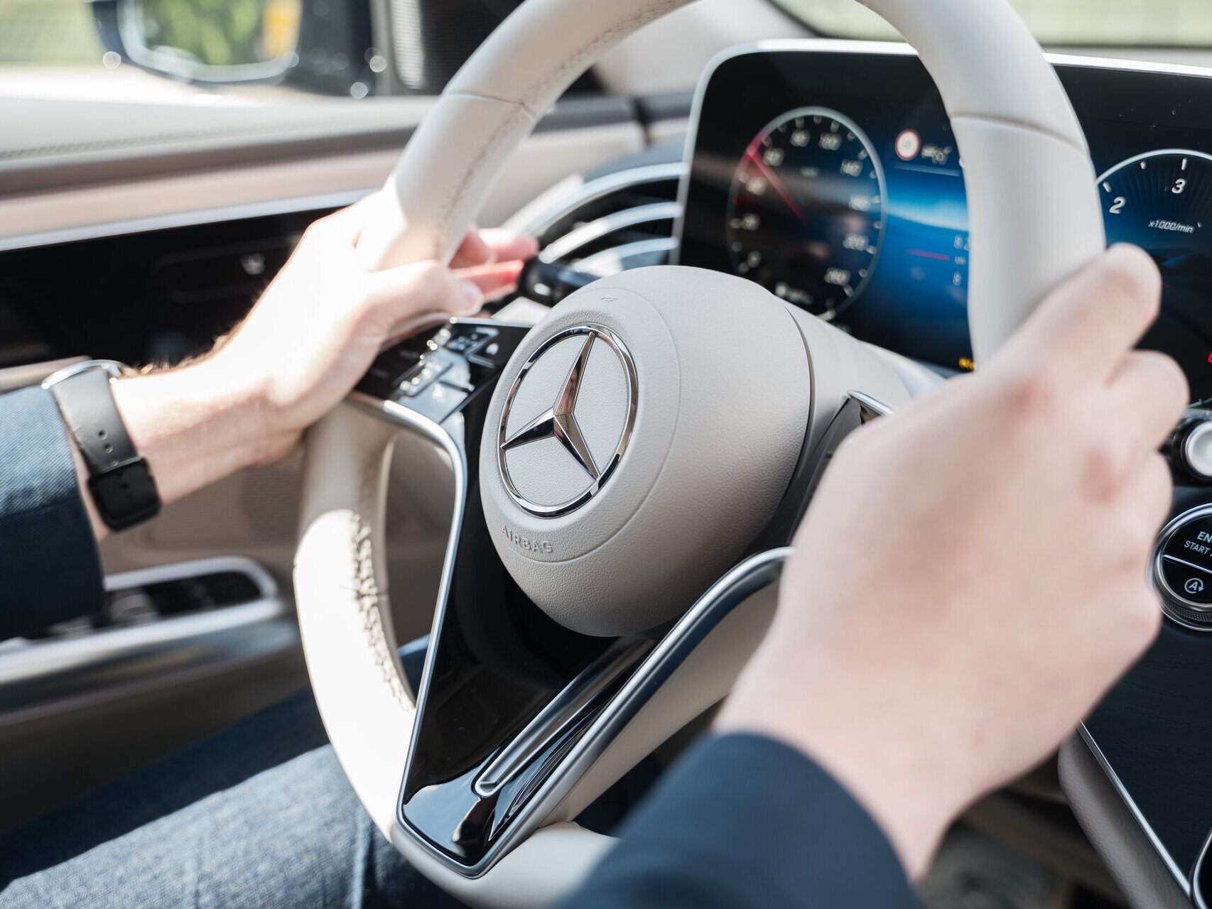 Hände eines Fahrers halten das Lenkrad eines Mercedes-Benz mit einem runden, silbernen Logo in der Mitte. Das Armaturenbrett ist im Hintergrund sichtbar.