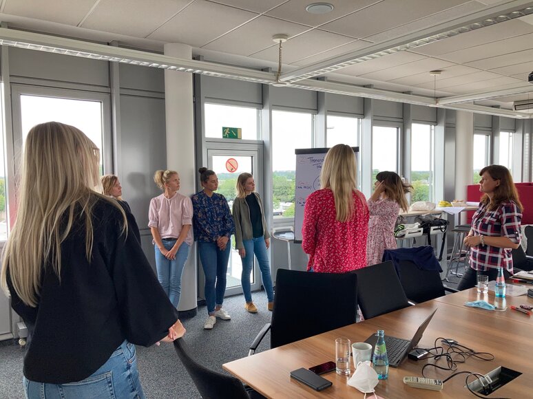 Eine Gruppe von sieben Frauen steht in einem modernen Büroraum, während eine Präsentation an einem Flipchart durchgeführt wird. Tische mit Büromaterialien sind im Vordergrund sichtbar, und durch große Fenster ist der Blick nach draußen zu sehen.