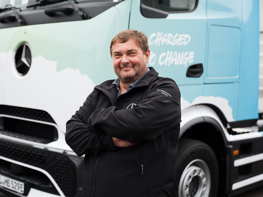 Ein Mann mit kurzen Haaren und einem Bart, der eine schwarze Jacke trägt, steht mit verschränkten Armen vor einem blau-weißen Mercedes-Benz-Lkw. Auf der Seite des Lkw steht "Charged to Change".