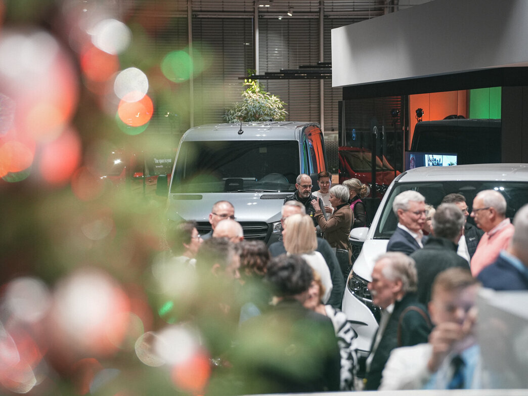 Eine Menschenmenge versammelt sich in einem Innenraum in der Nähe von weißen Fahrzeugen, möglicherweise auf einer Veranstaltung oder in einem Ausstellungsraum, wobei ein geschmückter Weihnachtsbaum im Vordergrund verschwimmt.