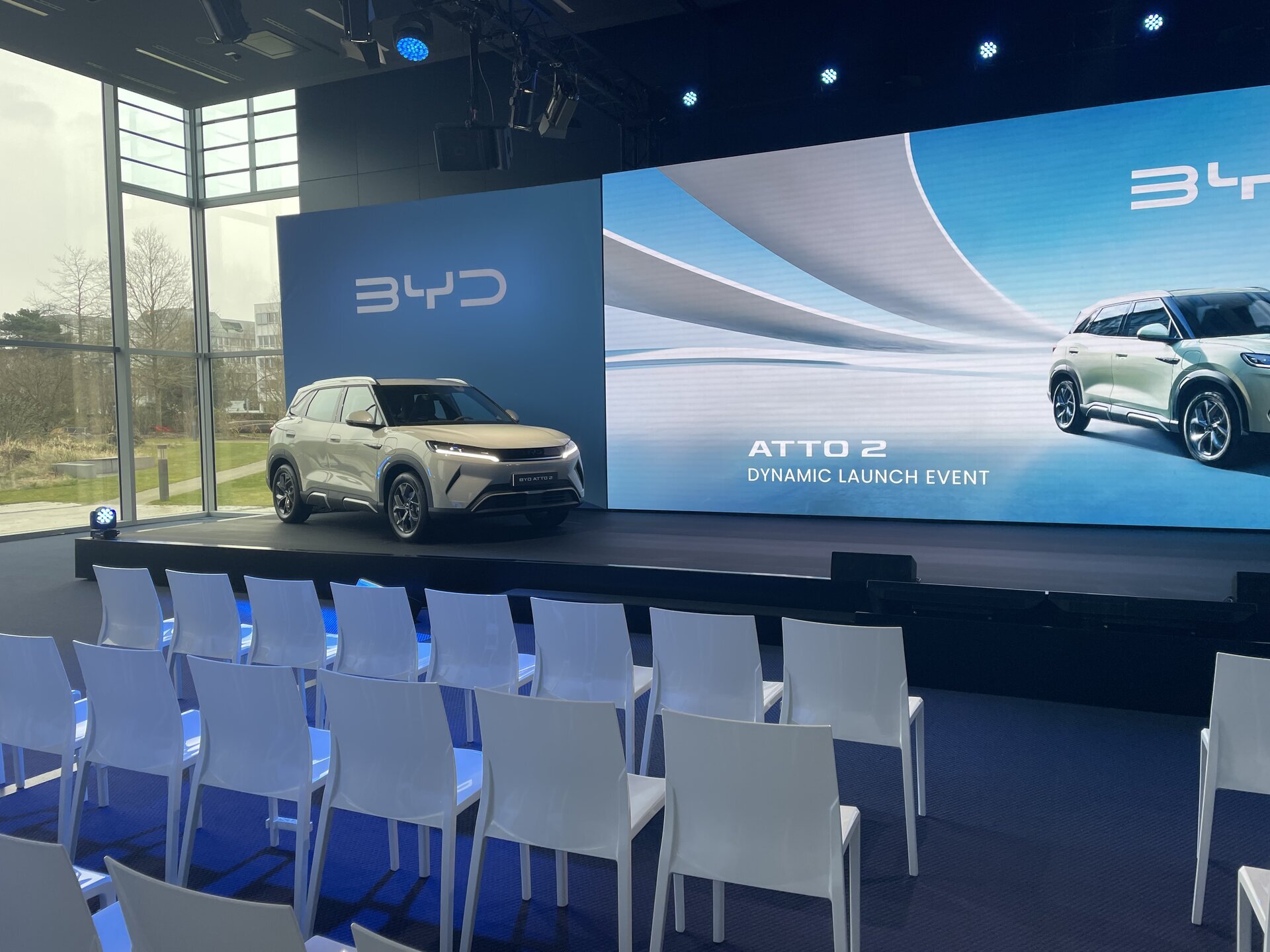 Moderner Präsentationsraum mit weißem BYD-Auto vor großer Leinwand, auf der 'ATTO 2 DYNAMIC LAUNCH EVENT' steht, und mehreren weißen Stühlen.