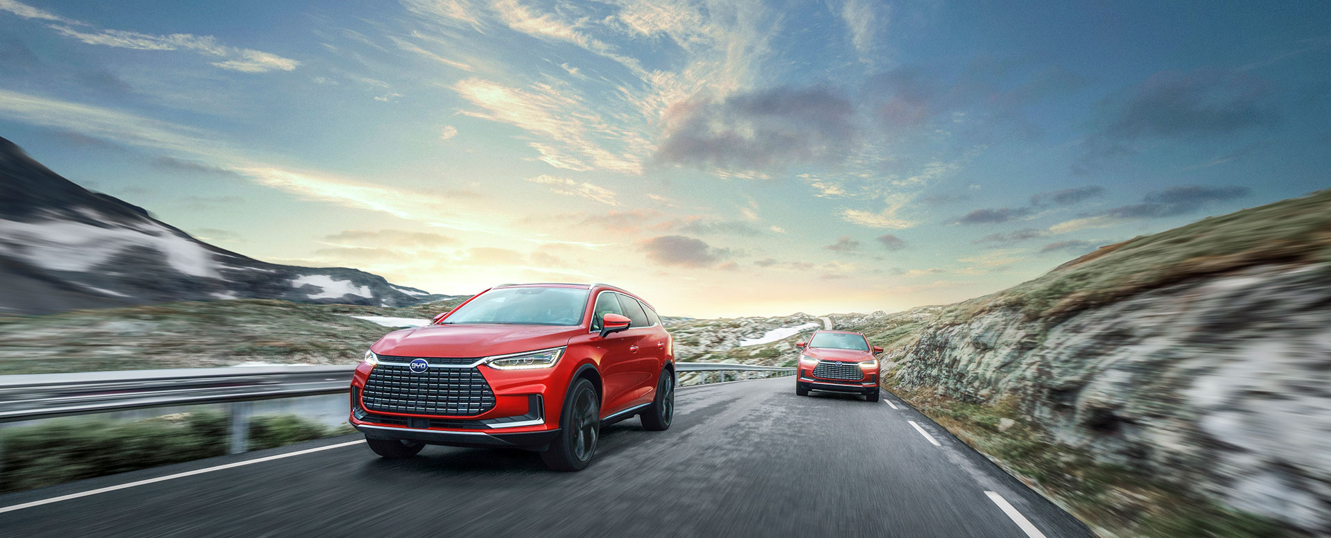 Zwei SUVs fahren auf einer kurvenreichen Straße mit einer malerischen Landschaft im Hintergrund. Der Vordergrund zeigt einen roten SUV, während der zweite SUV in der Entfernung zu sehen ist. Der Himmel ist klar mit sanften Wolken, und die Umgebung wirkt natürlich.
