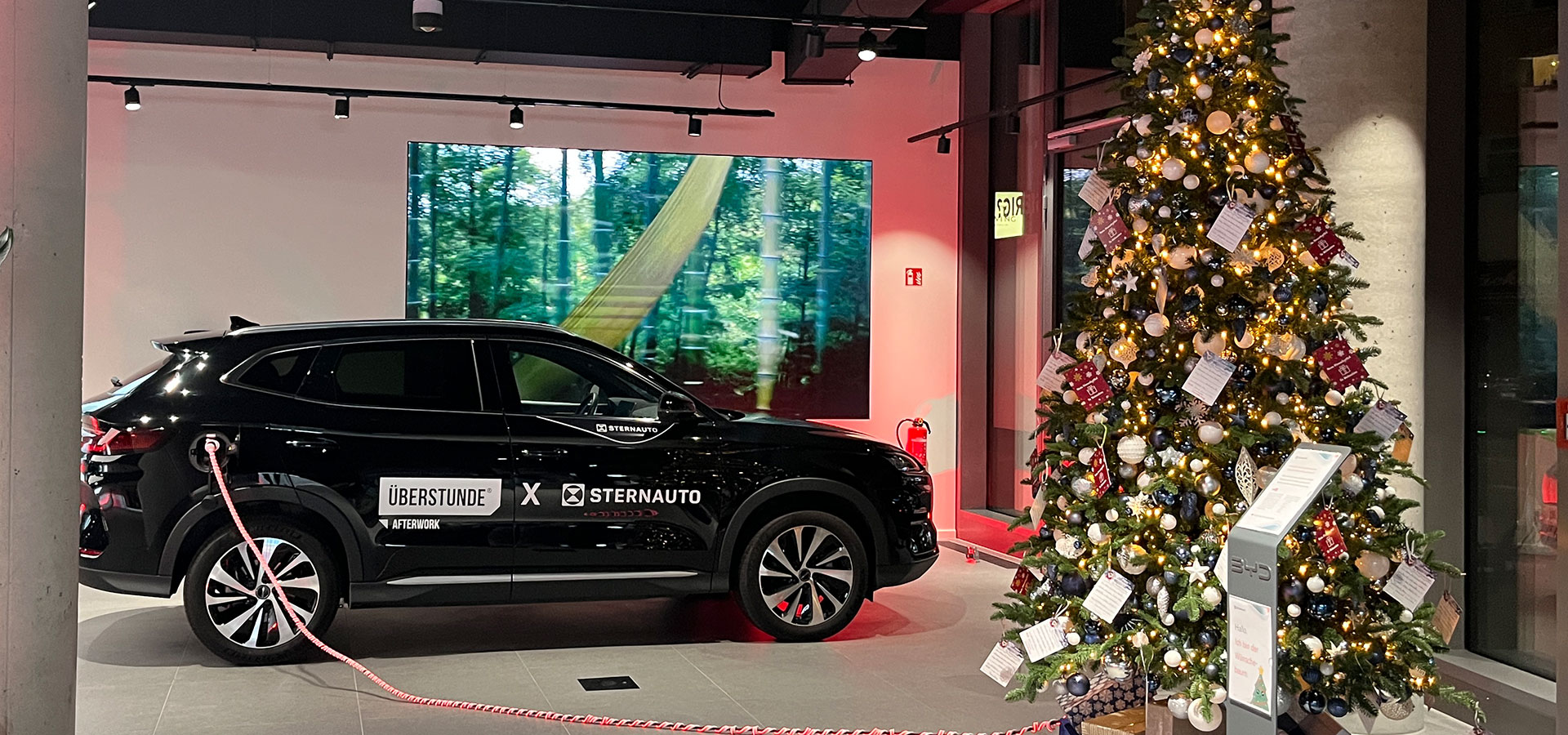 Ein schwarzes Auto ist in einem Innenraum neben einem großen, geschmückten Weihnachtsbaum geparkt. Das Auto ist an eine Ladestation angeschlossen, und an der Wand im Hintergrund ist ein Waldbild zu sehen.